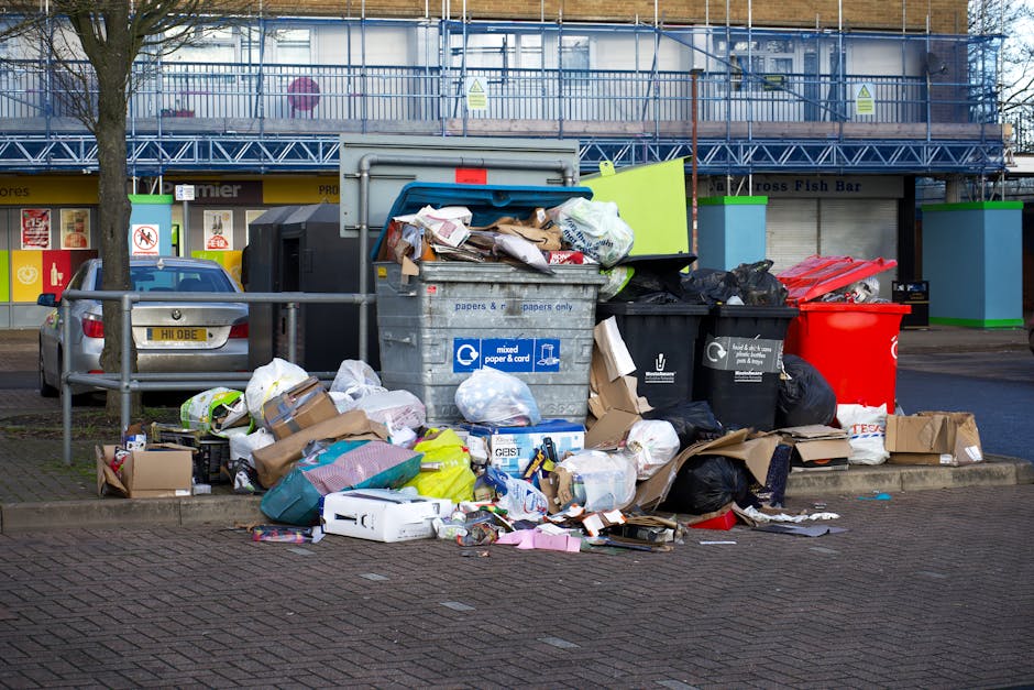 A large pile of mixed household waste and discarded packaging is accumulated on a paved urban sidewalk, with cardboard boxes, plastic bags, paper, and packaging materials spilling out of a gray recycling bin labeled for mixed paper and cardboard. Surrounding the bin are additional black and red rubbish bags, some partially torn, along with loose items like a small cardboard box and plastic containers scattered on the ground. On the left, a silver car is parked parallel to a metal railing that separates the sidewalk from the parking area. In the background, a building with large glass windows and a blue scaffold structure suggests under-construction or renovation work, with storefronts visible on the ground floor, including signage for a fish bar. The scene is illuminated by natural daylight, highlighting the cluttered and unmanaged appearance of the waste outside typical council collection zones, reflecting an instance of private rubbish handling or on-site clearance by Waste Disposal Pimlico.