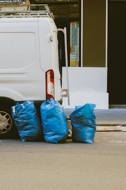 In this image, three large blue plastic waste bags are positioned on the pavement beside a white commercial van, which is partly visible with the rear door closed and the back wheel prominent on the left side. The bags appear to be filled with rubbish and are gathered close together, resting on a street surface that is smooth and grey. Behind the bags, part of a building is visible, featuring a large glass window or door with a dark frame and partial reflections, indicating an urban or commercial environment. The scene is evenly lit, suggesting natural daylight, and appears to depict a typical setting for private waste collection or rubbish removal, possibly in the context of a quick, independent collection service such as those offered by Waste Disposal Pimlico. The overall image reflects a straightforward scenario of waste disposal, with an emphasis on the handling of rubbish bags ready for collection in a street-based environment.
