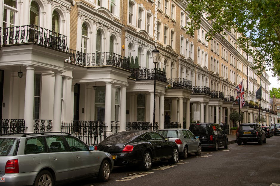 A row of white, multi-storey Victorian-style terraced houses with ornate architectural details, black wrought-iron balconies, and white columns supporting the porches along a street in Pimlico. Several parked cars, including a silver estate, a black coupe, a grey hatchback, and other vehicles, are positioned along the curb. In the background, a lamppost and British flags hang from the building facades. Green leafy trees extend over the scene, providing partial canopy cover. The street appears clean and well-maintained, with a quiet residential atmosphere typical of central London neighborhoods, illustrating a setting where private waste collection or rubbish removal services could be relevant. Waste Disposal Pimlico may provide services to maintain this tidy appearance, especially during estate clearance or routine rubbish removal, supporting alternative waste handling options outside municipal collection. The lighting is natural daylight, with soft shadows cast by the trees and buildings, emphasizing a clear and calm environment suitable for professional rubbish disposal services.