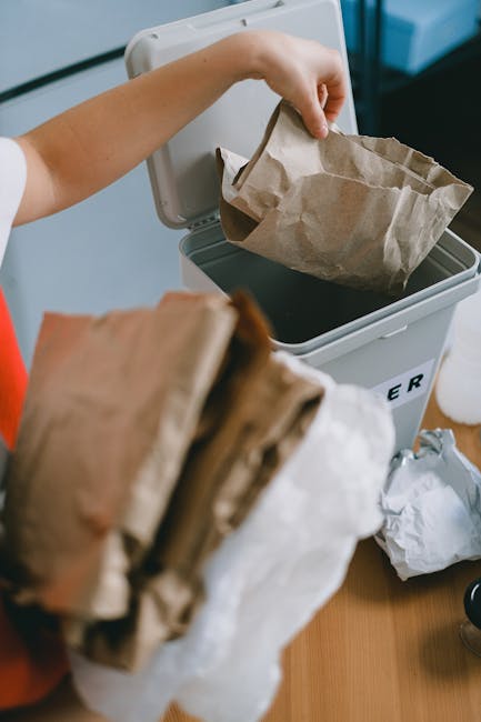 A person is seen placing a crumpled brown paper bag into a grey rubbish bin with a partially open lid, situated indoors on a wooden surface. The paper bag appears to be filled with lightweight waste and has a slightly textured surface. In the foreground, another brown paper bag is being held, showing its crumpled and folded edges. Beside the bin, there are additional crumpled paper bags and waste materials, indicating an active process of rubbish collection or disposal. The environment suggests a domestic setting, with a focus on waste management, possibly relating to household rubbish removal services offered by Waste Disposal Pimlico. The scene emphasizes the handling of reusable or biodegradable waste, aligning with best practices for environmentally conscious waste disposal and private collection options that do not involve local authority services. The overall lighting is natural and soft, ensuring the details of the paper materials and the process are clearly visible and easy to interpret for accessibility purposes.