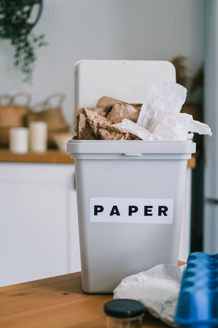 A grey paper recycling bin placed on a wooden surface indoors, filled with crumpled paper, torn cardboard, and paper packaging, with some papers protruding from the top. The bin has a white label with the word 'PAPER' in black capital letters. In the background, there are blurred household items including candles and wicker baskets on a surface, and a green potted plant hanging from the ceiling. The environment suggests an organized home setting suitable for waste sorting and waste management practices, supporting the principles of household waste separation and independent collection services like those offered by Waste Disposal Pimlico.