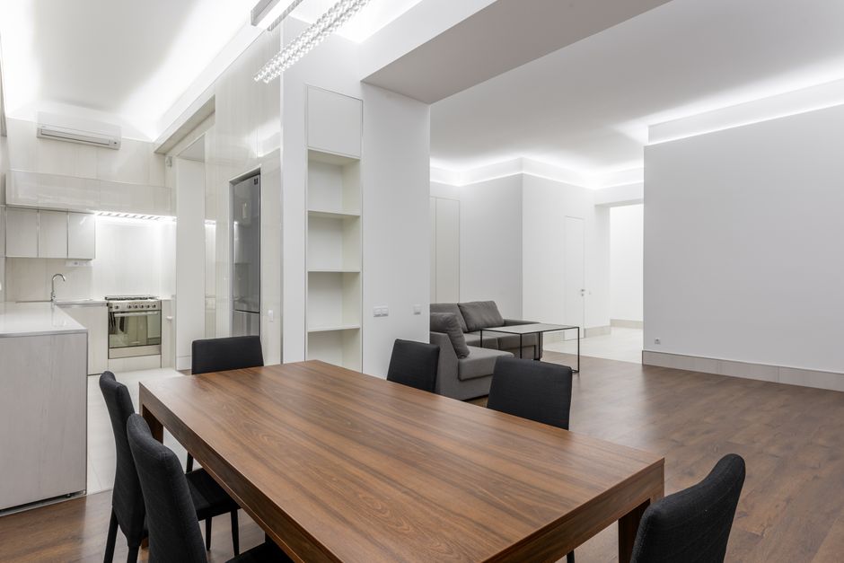 A spacious, modern interior featuring a dining area and living space in an apartment or house with minimalist design. The foreground shows a rectangular wooden dining table with a smooth, polished surface and six dark upholstered chairs arranged around it. Behind the dining area, there is a kitchen with a white countertop, light-colored cabinets, and a stainless steel oven and refrigerator, all set against a tiled wall. To the right, a sitting area includes a gray fabric sofa with cushions situated near a low rectangular coffee table. The room is brightly lit by natural light coming from a ceiling window or skylight, with white walls and ceiling creating a clean, contemporary ambiance. The floor is covered with medium-tone wood laminate. The overall environment appears tidy and uncluttered, with no visible rubbish or waste, reflecting a well-maintained living space, possibly prepared for a professional rubbish removal or interior cleaning service by Waste Disposal Pimlico.