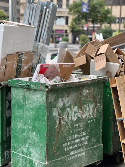 A worker wearing a light blue shirt, dark trousers, gloves, and a white face mask is standing on the back of a large black waste collection truck in an urban environment. The worker is using a long-handled tool to load or rearrange a variety of discarded items, including wooden panels with different finishes—some smooth, polished surfaces in brown and white tones, others with a more weathered appearance. The truck is parked on a paved surface with a building featuring modern glass and concrete architecture in the background. The truck's cargo area is filled with waste materials typical for private or alternative rubbish collection, with some items leaning or stacked upright. Natural daylight illuminates the scene, highlighting the textures of the wood, the metal truck, and the surrounding cityscape, subtly illustrating a professional waste removal activity suitable for independent rubbish disposal services like Waste Disposal Pimlico.
