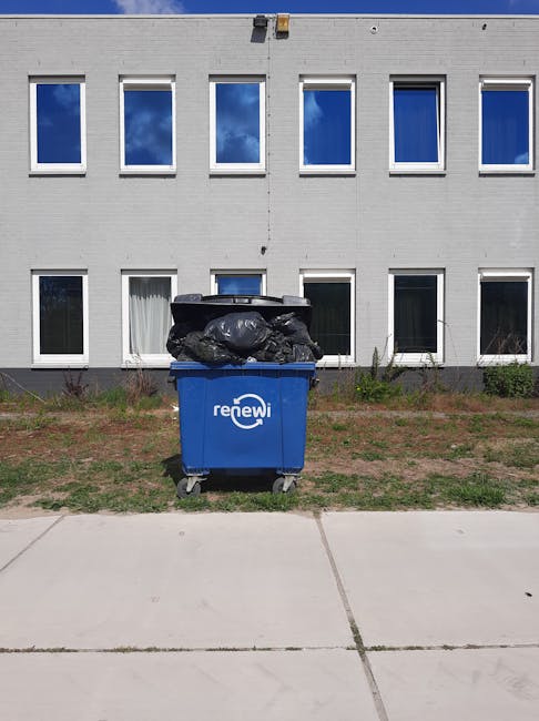 A blue wheeled trash bin with the branding 'renewi' is positioned on a concrete sidewalk in front of a modern, white-painted building with large rectangular windows. The bin is filled with black rubbish bags stacked on top of each other, some of which appear to be slightly bulging or loosely tied. The area surrounding the bin features a narrow strip of grass and small bushes or plants directly behind it, indicating an outdoor urban environment. The building's exterior is smooth with evenly spaced framed windows reflecting the blue sky and some clouds, suggesting a bright, clear day. The scene emphasizes the presence of a public waste collection point, which could relate to private on-site or neighbourhood rubbish disposal services, aligning with the context of commercial waste removal and alternative rubbish collection methods supported by Waste Disposal Pimlico.