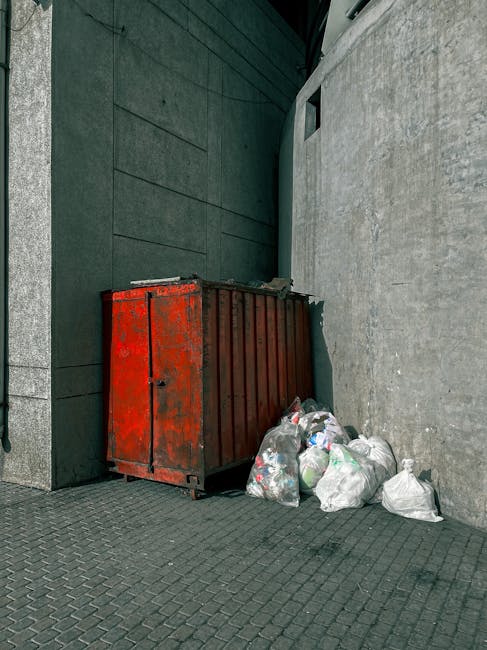 A weathered, red metal skip positioned against a dark green wall on a paved urban street. The skip has a rectangular shape with vertical ridges along its sides and visible signs of rust and peeling paint, indicating frequent use. Next to it, on the pavement, there are several tightly tied white plastic rubbish bags filled with mixed waste, including visible plastic bottles and other debris. The bags are stacked haphazardly, some leaning against the skip and others resting on the uneven, textured stone surface. To the right, a plain, light grey concrete wall extends into the background, with a small, narrow window or vent situated high on it. The surrounding environment appears quiet, with no other objects or pedestrians visible, suggesting this scene is typical of a private or independent waste collection point in an urban setting, possibly managed by a specialist waste disposal service like Waste Disposal Pimlico, supporting alternative rubbish removal methods apart from council collection.