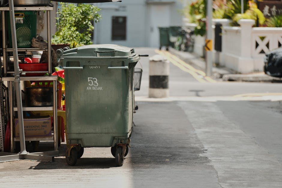 A large green wheeled waste bin with a closed lid positioned on the sidewalk in front of storage shelves holding various household items and tools. The bin has a weathered appearance with visible scuff marks and white lettering indicating the number 53 and the words 'JUN BESAR'. It is situated next to a metal shelving unit containing jars, boxes, and other miscellaneous objects, with part of a red and yellow container visible on the same shelf. The scene is outdoors on a paved street with a driveway, yellow road markings, and a white stone wall with black and yellow safety barriers in the background. The lighting suggests bright daytime, with natural sunlight casting shadows. This setup is typical of private waste collection or rubbish removal services operating on urban streets, with Waste Disposal Pimlico providing such services for waste management and alternative disposal purposes in urban environments.