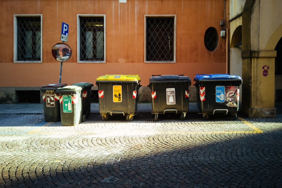 A weathered, red metal skip positioned against a dark green wall on a paved urban street. The skip has a rectangular shape with vertical ridges along its sides and visible signs of rust and peeling paint, indicating frequent use. Next to it, on the pavement, there are several tightly tied white plastic rubbish bags filled with mixed waste, including visible plastic bottles and other debris. The bags are stacked haphazardly, some leaning against the skip and others resting on the uneven, textured stone surface. To the right, a plain, light grey concrete wall extends into the background, with a small, narrow window or vent situated high on it. The surrounding environment appears quiet, with no other objects or pedestrians visible, suggesting this scene is typical of a private or independent waste collection point in an urban setting, possibly managed by a specialist waste disposal service like Waste Disposal Pimlico, supporting alternative rubbish removal methods apart from council collection.