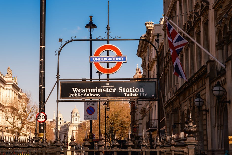 A detailed street scene in Westminster featuring the iconic London Underground sign with the red circle and blue bar, positioned above a rectangular black signboard indicating Westminster Station, alongside directions for the Public Subway and Toilets. To the right, a Union Jack flag is mounted on a pole attached to a historic building with ornate architecture and arched windows. In the background, classic Westminster-style architecture is visible, including a white dome-topped structure, set against a clear blue sky. Foreground elements include a black lamppost with decorative scrollwork, a street sign indicating a 20 mph speed limit, and a small blue no-parking zone sign. The scene is illuminated by natural daylight, highlighting the textures of the stone facades and metallic fixtures. The overall atmosphere captures the vibrant and organized urban environment typical of central Westminster, with a subtle emphasis on the area's transport infrastructure that relates to alternative waste management solutions, often serviced by private rubbish removal companies like Waste Disposal Pimlico.